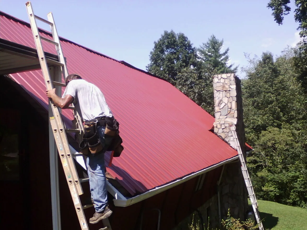 Roofer on a ladder beside a red metal roof and stone chimney — Soffit & Fascia Repair by Roofing Brothers in Fergus Falls